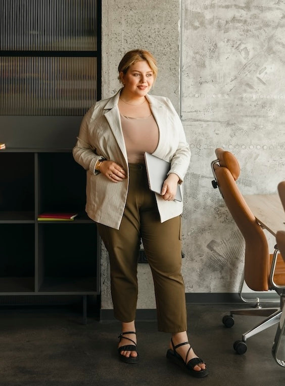 Une femme ronde confiante au bureau portant une veste ouverte claire, un pantalon kaki et un ordinateur portable.